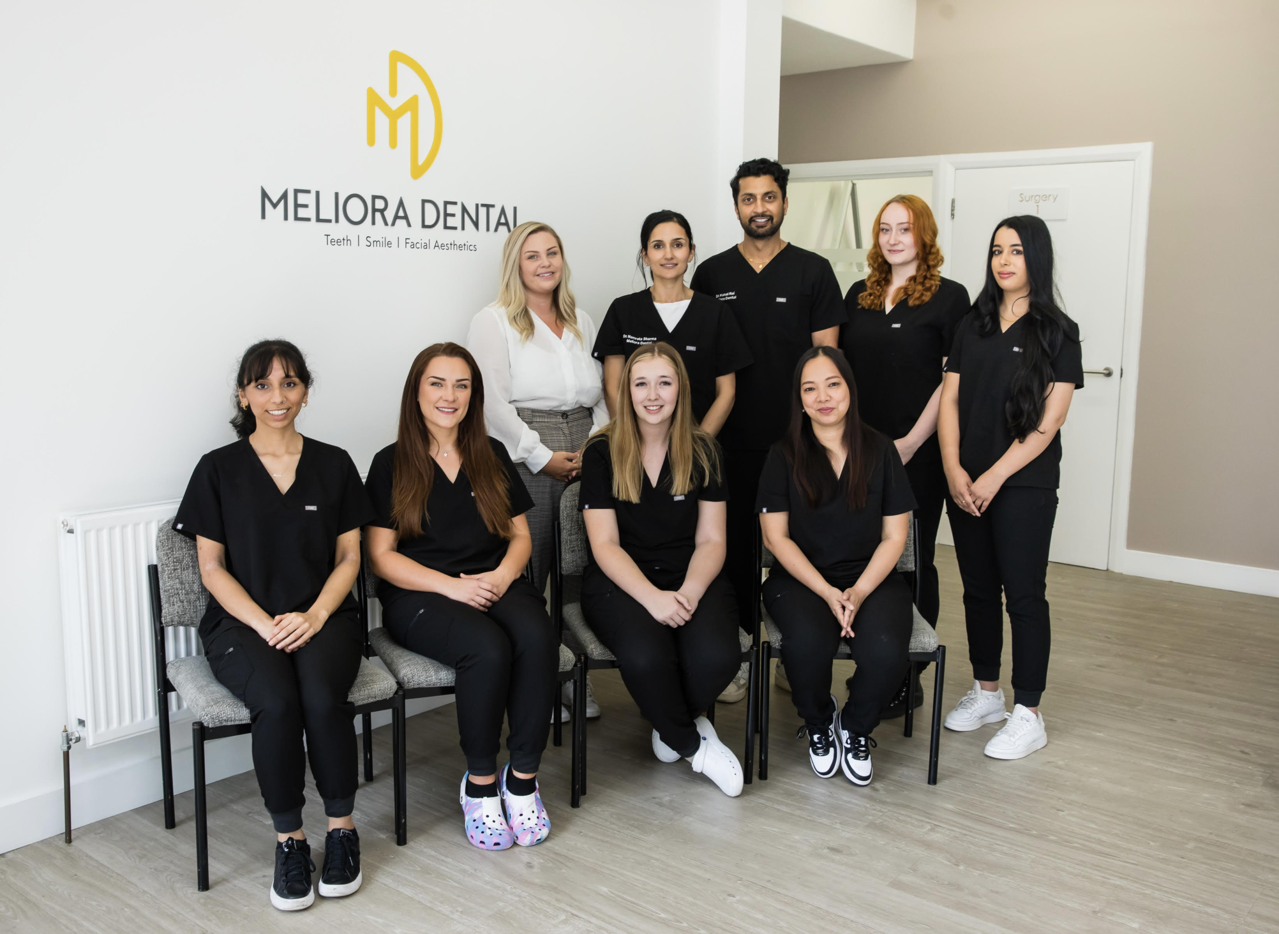 1 male and 8 female employees of a dental practice. Neutral walls. Meliora Dental logo is printed on the wall. 